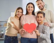 Happy mother father and two children posing together showing big heart postcard celebrating Valentines Day in family circle, have good relationships express love to everyone, posing over home interior
