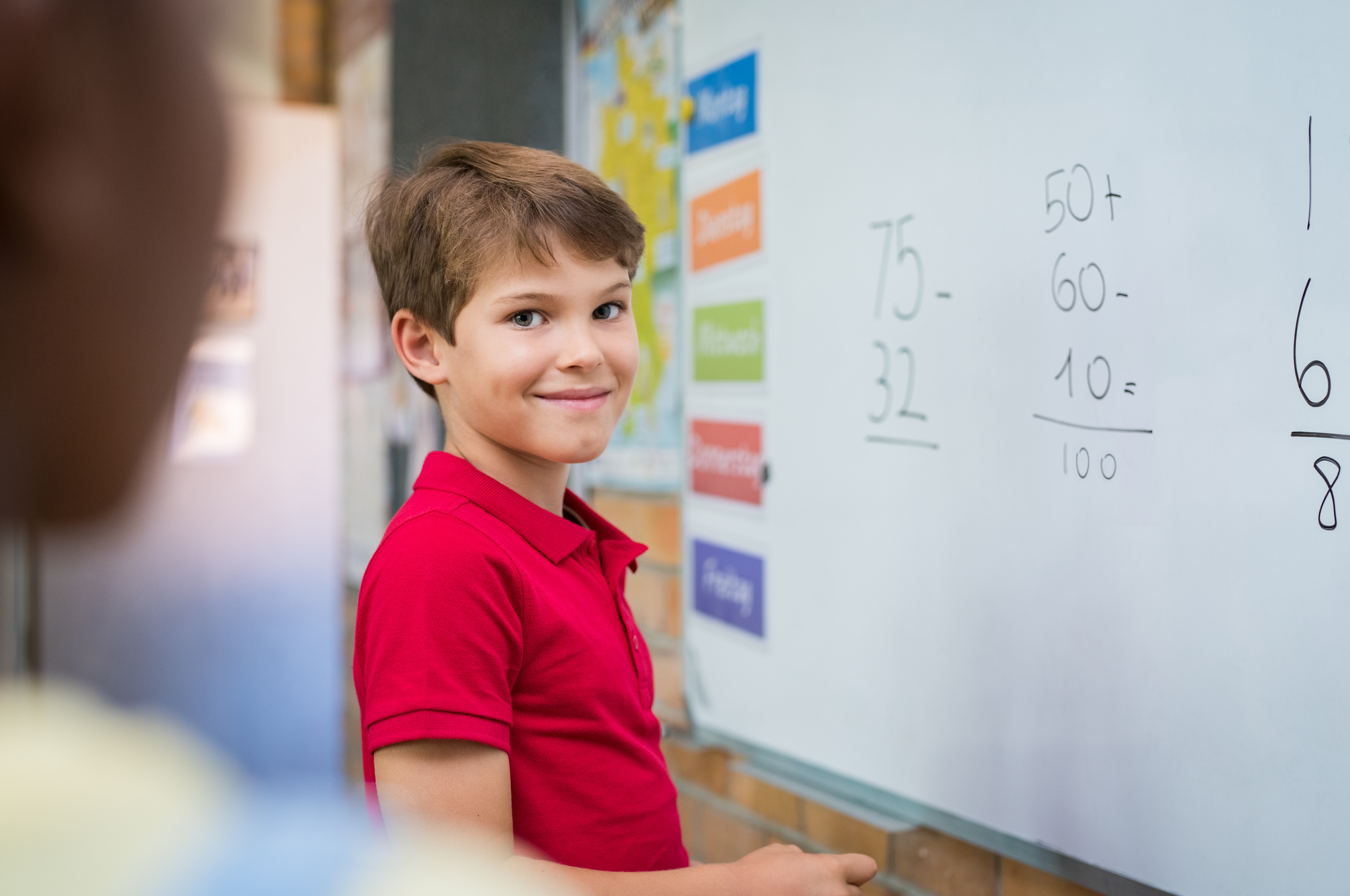 Clever boy doing sums on white board and looking at camera. Smiling schoolboy solving addition and subtraction sum in mathematics lesson. Proud young boy standing with math operation solved.