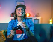 Handsome Excited Young Gamer with Long Hair and a Cap is Sitting on a Couch and Playing Video Games on a Console. He Plays with a Wireless Controller. Cozy Room is Lit with Warm and Neon Light.