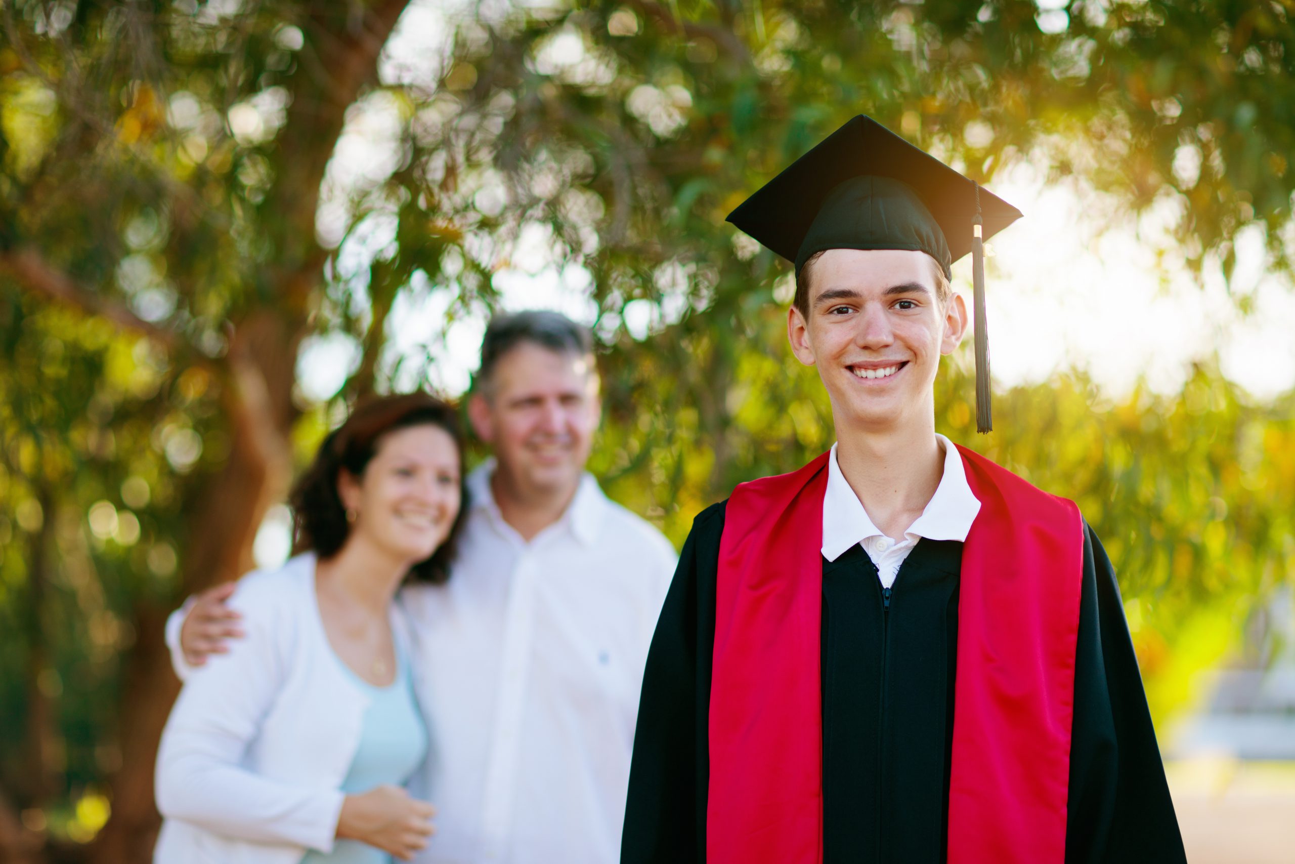 School or college graduation ceremony. Young man in gown and cap, with his family. Moher and father cheer at celebration of successful diploma certificate. High school graduate in robe and mortarboard