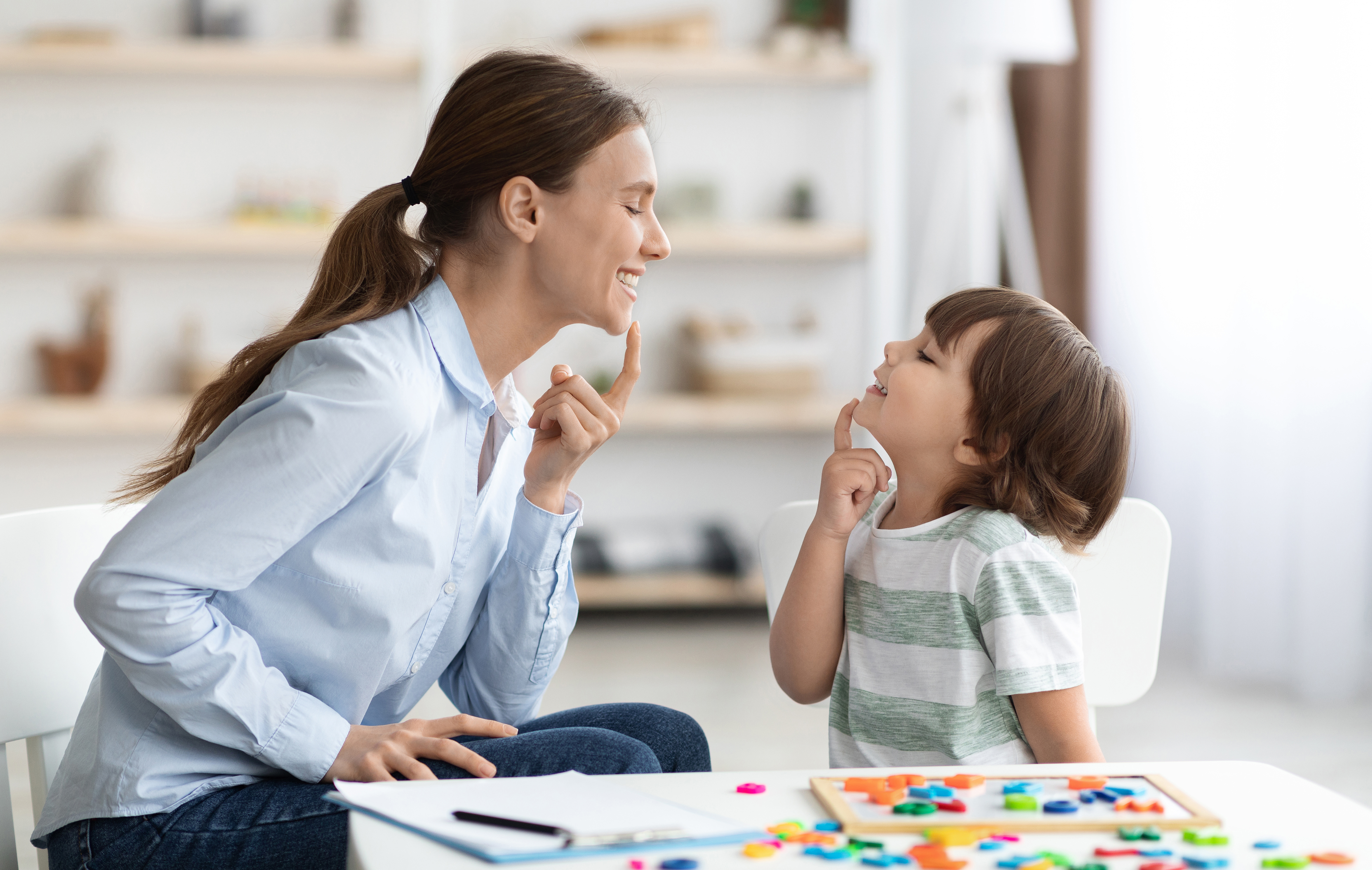 Speech training for kids. Professional woman specialist training with little boy at cabinet, teaching him right articulation exercises, side view