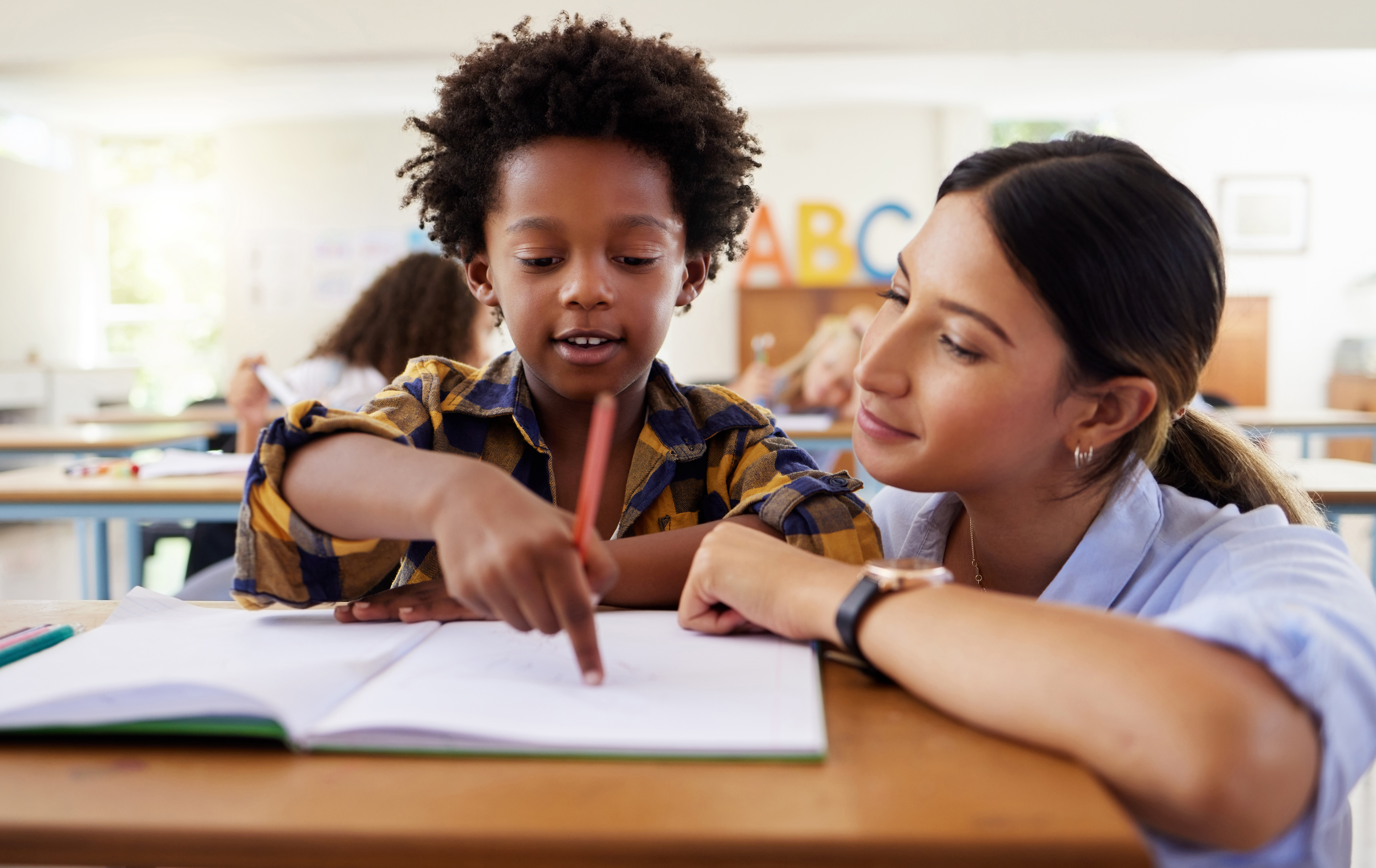 Teacher, learning and helping black kid in classroom for knowledge, studying or assessment. Question, development and boy or student with woman for education pointing in notebook in kindergarten